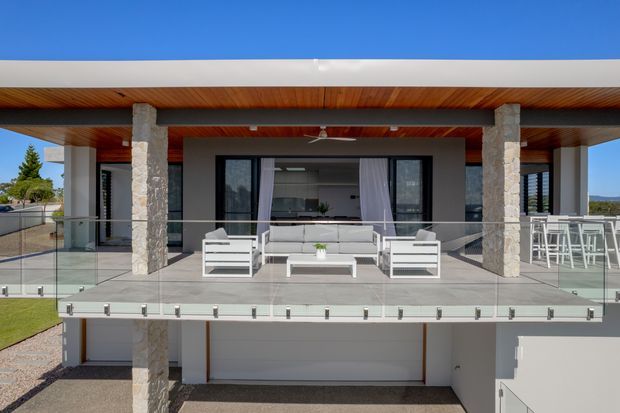 Modern two-storey home exterior viewed from the front, featuring a broad flat roof, timber-lined soffits, stone-clad columns, extensive black-framed glazing, and a glass-balustraded upper-level terrace furnished as an outdoor lounge. A garage is visible below the terrace.