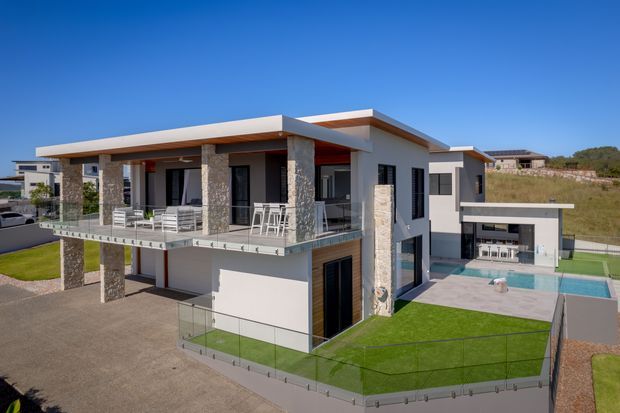 A contemporary multi-level residence viewed from the exterior, featuring flat roof planes, large glazed openings, stone-clad structural columns, white rendered walls, broad balconies with glass balustrades, and a private swimming pool terrace. The home sits in a suburban development with landscaped lawn and open surrounding terrain.