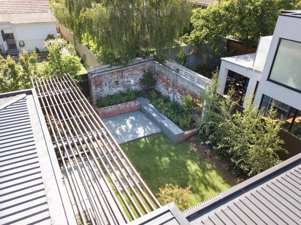 Elevated view over a landscaped residential courtyard enclosed by a weathered brick boundary wall. The garden includes a lawn, mature trees and planting, a paved or concrete seating platform with a low built-in bench, and a prominent timber pergola with slatted screening beside corrugated metal roofing. Modern white residential architecture frames the courtyard on the right, with neighbouring homes visible beyond.