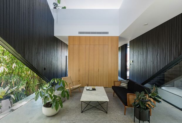 A contemporary double-height living room with a dramatic central skylight and warm timber feature wall. Dark vertical wall panelling runs along both sides, with full-height glazing and greenery to the left. A neutral sofa, leather lounge chairs, pale rectangular coffee table, indoor plants, and a staircase create a refined, natural interior.