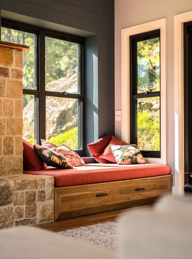 A warm, sunlit interior nook with a built-in window seat beneath tall black-framed windows. The seat has a deep red cushion and colourful patterned pillows, with substantial timber drawer storage below. A rustic exposed-stone masonry feature frames the left side, while dark painted walls and white-trimmed openings create contrast. Natural landscape is visible through the glazing.