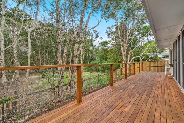 A spacious timber deck extending along the side of a home, with warm-toned timber decking and railing, dark wire balustrades, and a covered roof edge. The deck overlooks a leafy natural landscape with mature trees and a fenced boundary. Glazed doors connect the deck to the interior of the house.