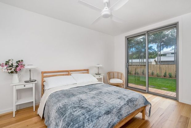 Bright, minimalist bedroom with a timber bed frame and matching light timber floor, white walls and bedding, simple white bedside tables, a woven accent chair, ceiling fan, and large sliding glass doors opening to a fenced lawn.