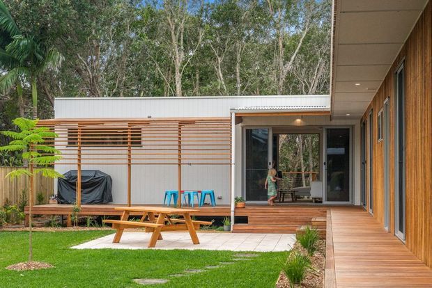 A contemporary single-storey dwelling arranged around a landscaped courtyard. The image shows a corrugated light-metal-clad pavilion with horizontal timber screening, extensive timber decking, large glazed sliding doors, and a covered outdoor dining area with a picnic table. A second timber-clad wing frames the right side, with lawn, paving, and established vegetation surrounding the home.