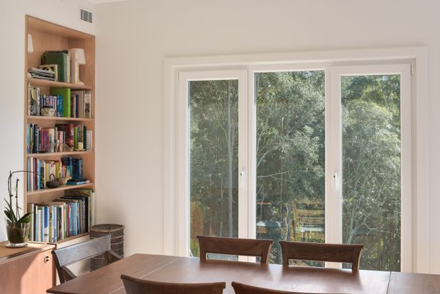 A bright, compact dining area with a rectangular timber dining table and chairs positioned in front of tall white-framed windows. Built-in timber shelving on the left is filled with books and small objects, with a potted plant and low timber cabinetry nearby. Dense greenery is visible outside through the glazing.