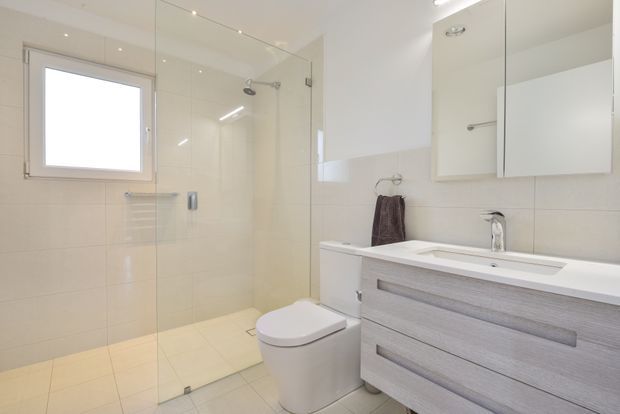 Bright, contemporary bathroom with a frameless glass walk-in shower, wall-hung toilet, long double vanity with pale grey wood-look drawers, white integrated basins, mirrored wall cabinets, and large light beige tiles. A small frosted window provides natural light, while recessed ceiling lighting keeps the room clean and evenly illuminated.