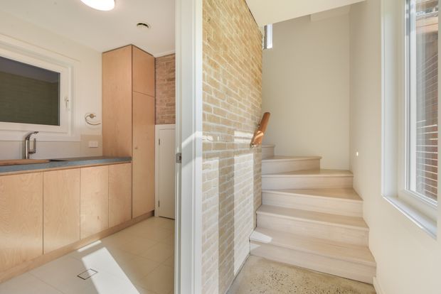 Bright interior view showing a compact kitchen on the left and a stairway rising beside an exposed pale brick wall. The kitchen has light timber cabinetry, a dark benchtop, tiled flooring, a sink, and integrated appliances. The stairs have light timber or timber-look treads, a timber handrail, and a large white-framed window providing abundant natural light. The overall design is clean, warm, and contemporary with a restrained material palette.