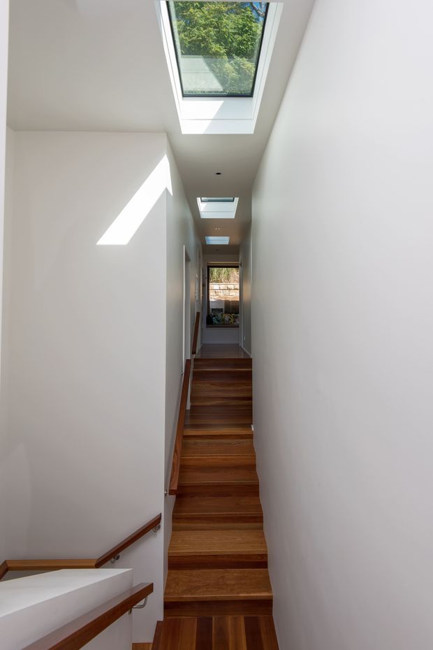 A narrow, bright interior stairway and hallway with a long flight of dark timber stairs, white plastered walls, timber handrails, and multiple rectangular skylights bringing in natural light. The stairway rises toward a small landing and window at the far end.