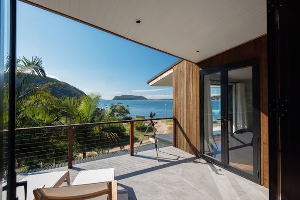 A bright covered outdoor terrace overlooking a tropical coastal bay. The terrace features pale tiled flooring, a dark metal-and-timber cable balustrade, timber-clad walls, a white paneled soffit with recessed downlights, and large black-framed glazed doors opening into the interior. Palm foliage, wooded hills, blue water, and a distant headland are visible beyond the deck.