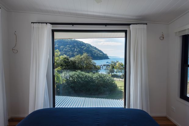 A bright bedroom viewed toward a large black-framed window with white curtains, overlooking a lush coastal landscape, hillside, and blue water. The room has a white painted wall and beadboard-style ceiling, timber flooring, and a bed dressed in dark navy bedding. A second black-framed window is visible on the right.