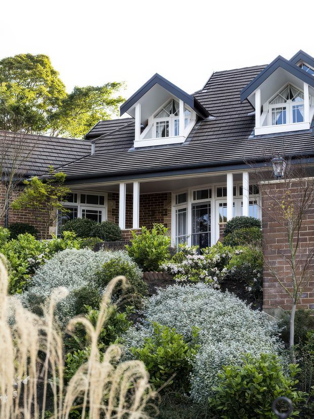 Exterior view of a substantial brick house with a complex dark tiled roof, multiple white-trimmed gabled dormer windows, covered verandah, and landscaped front garden. The facade combines dark brown brick, white architectural trim, large divided windows, and layered pitched roof forms.