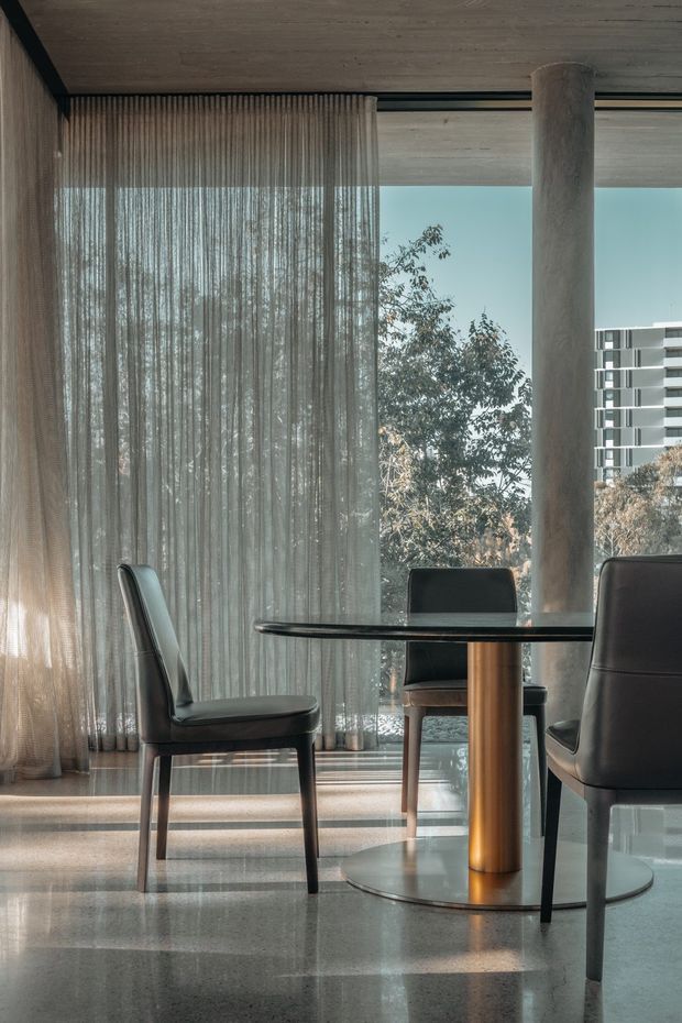 A refined dining area with a round dark tabletop supported by a cylindrical timber pedestal and glass base, surrounded by upholstered dining chairs. Full-height sheer curtains frame expansive glazing, with a concrete column and polished concrete or terrazzo-like floor creating a calm, modern interior. Trees and a neighbouring apartment building are visible outside.