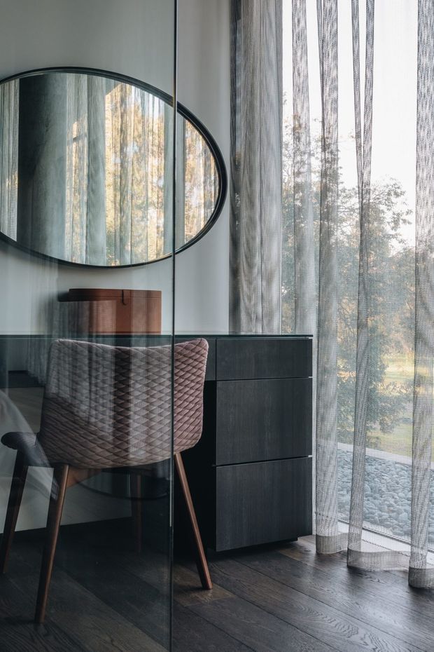 A refined home office or dressing-area workspace beside floor-to-ceiling sheer curtains. The room features a dark timber desk and matching tall drawer cabinet, a textured upholstered chair with timber legs, a large oval black-framed wall mirror, and dark timber flooring. The glazing overlooks a landscaped outdoor setting and creates soft, filtered natural light. The image has a calm, sophisticated, contemporary feel.