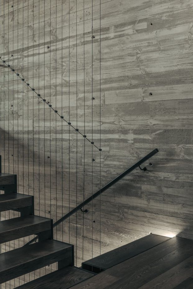 Close-up architectural photograph of a contemporary stairway with dark timber treads and landing, a slim black metal handrail, and a dramatic full-height exposed concrete wall. Vertical suspension cables with small circular fittings run beside the stair, creating a graphic linear pattern. The moody, industrial material palette is highlighted by subtle recessed lighting.