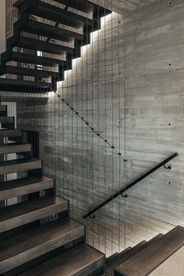 Interior architectural photograph of a dramatic stairway with open-riser dark timber treads, black metal stringers and handrail, and a transparent cable balustrade. The stairs run alongside a large exposed concrete wall, creating a tall, sculptural circulation space with strong vertical and diagonal lines. The palette is dark, industrial, and refined.