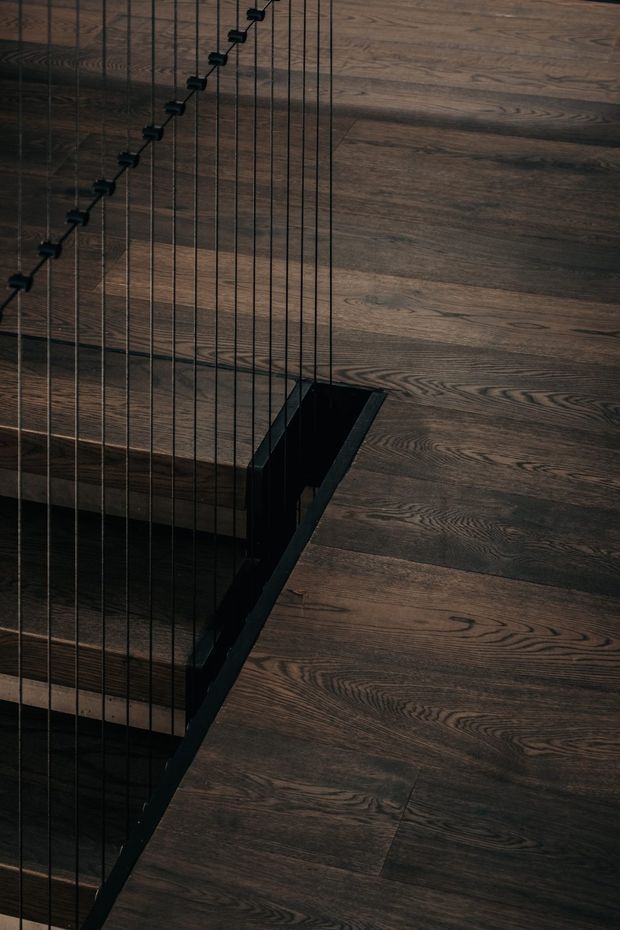Close-up architectural detail of a dark timber staircase and landing. Deep brown wood flooring and stair treads are paired with a black metal stair edge and slender vertical cable balustrade, creating a moody, refined interior composition.