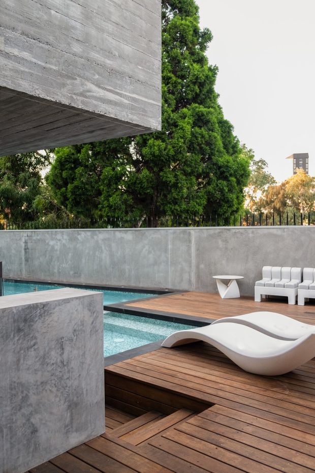 A contemporary outdoor pool courtyard with a long rectangular swimming pool, timber decking, rendered concrete privacy walls, white sculptural loungers, and outdoor seating. A projecting concrete building volume frames the upper left, while mature trees surround the space.