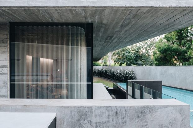 A contemporary concrete house exterior with a deep cantilevered concrete roof slab and matching rendered walls. A large dark-framed glazed opening with sheer curtains reveals the interior, while a narrow linear swimming pool and minimalist paved terrace extend alongside the building. Dense greenery is visible beyond the boundary wall.