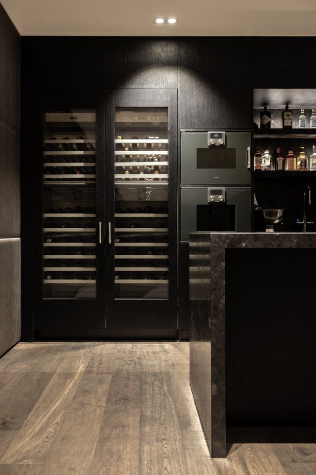 A dark, sophisticated home bar with floor-to-ceiling black timber cabinetry, two glass-fronted wine refrigerators, integrated appliances, open liquor shelving, and a substantial dark stone bar counter. Warm recessed lighting highlights the richly textured finishes, while wide timber flooring adds contrast and warmth.