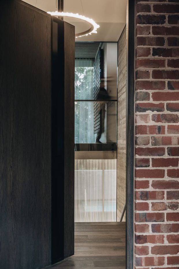 A narrow, dramatic interior passage framed by a dark timber pivot door or panel on the left and an exposed red brick wall on the right. The view leads toward a reflective glazed opening and a softly screened interior beyond, with dark timber flooring, warm neutral walls, and a sculptural circular pendant light. The space has a refined, moody, contemporary-industrial character with strong vertical lines and layered reflections.