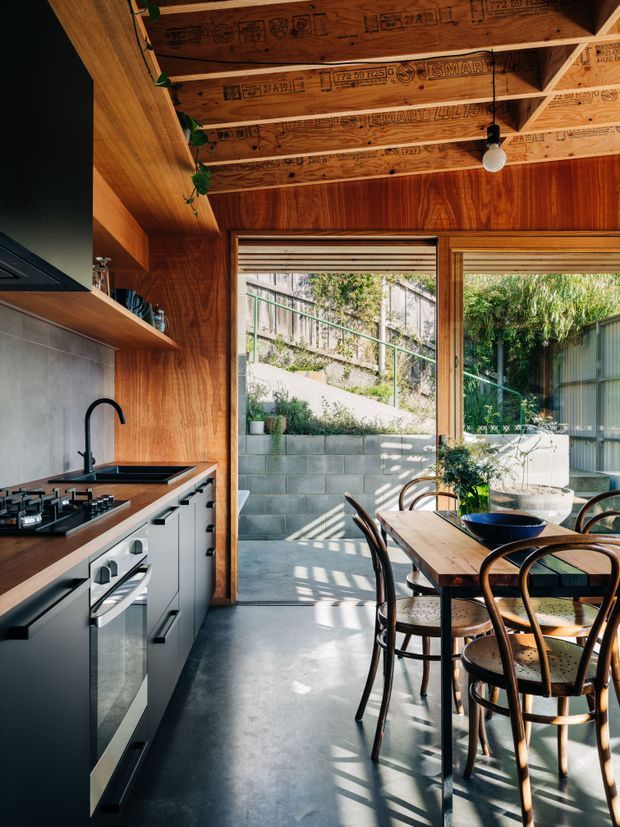 A compact open-plan kitchen and dining area with a warm timber-lined interior. The kitchen has white built-in cabinetry, dark benchtops, a tiled splashback, integrated cooking appliances and a black gooseneck tap. A timber dining table with curved wooden chairs sits beside large glazed openings leading to a small paved courtyard. The unfinished exposed timber roof framing and insulation markings are visible overhead, creating a strong contemporary workshop-like character. Sunlight casts patterned shadows across the polished grey concrete floor.