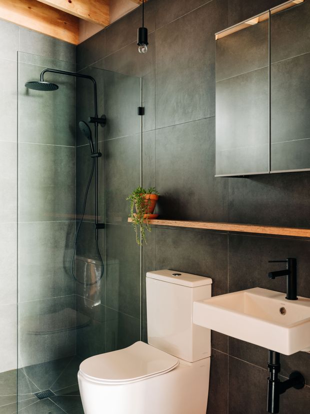 Compact modern bathroom with a walk-in glass shower, large dark grey wall tiles, black shower and tapware, a wall-mounted white basin, toilet, floating timber shelf, mirrored cabinet, and a warm exposed timber ceiling detail. A small potted plant adds greenery.