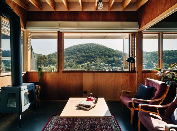 A warm, timber-lined living room with exposed ceiling beams, expansive corner glazing, and panoramic views of a densely vegetated hillside. The room features a freestanding black wood-burning stove, mid-century-style lounge chairs, a light rectangular coffee table, patterned rug, and indoor plants. The interior has rich dark timber surfaces and a strong connection to the surrounding landscape.