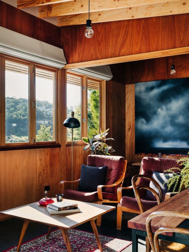 Warm, richly timber-lined living room with large wood-framed windows, exposed plywood ceiling beams, mid-century lounge chairs upholstered in burgundy, a low geometric coffee table, patterned rug, plants, and a large television. The room has a strong retro mid-century character with abundant natural light and views through the windows.