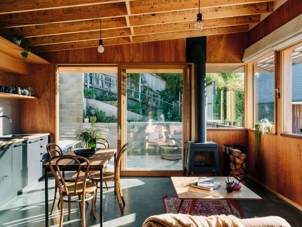 A compact, warm timber-lined open-plan kitchen and living area with exposed timber ceiling beams, large glazed doors and windows, a small dining table with bentwood chairs, compact cabinetry, and a freestanding wood-burning stove beside stacked firewood. Sunlight enters from the glazing and opens toward a planted, terraced courtyard or garden.