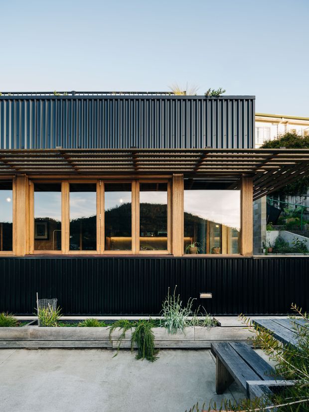 Exterior view of a contemporary residential building with a dark vertically ribbed metal-clad upper and lower facade, warm timber-framed glazing, and a broad timber pergola or brise-soleil. A landscaped paved courtyard with raised planter beds and timber seating occupies the foreground. The architecture has a strong linear composition and contrasts dark metal, natural timber, and concrete paving.