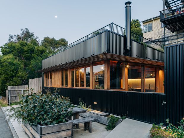 Exterior view of a compact contemporary dark-clad building with a warm timber-framed glazed addition. The structure has a rooftop terrace enclosed by vertical metal balustrades, a prominent metal chimney, extensive windows, and a small paved garden courtyard with raised planter boxes and bench seating.