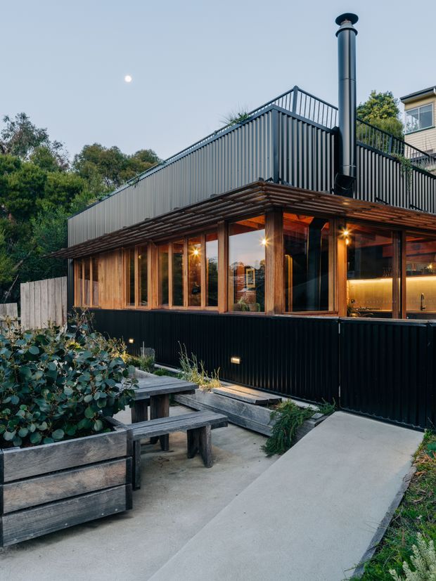 Exterior dusk view of a contemporary, multi-level home or extension with dark corrugated metal cladding, a low sloping roof, timber-framed glazing, and a prominent black flue. The building opens onto a landscaped concrete patio with raised timber garden beds and built-in bench seating. Warm interior lighting reveals a kitchen or bar area through the windows.