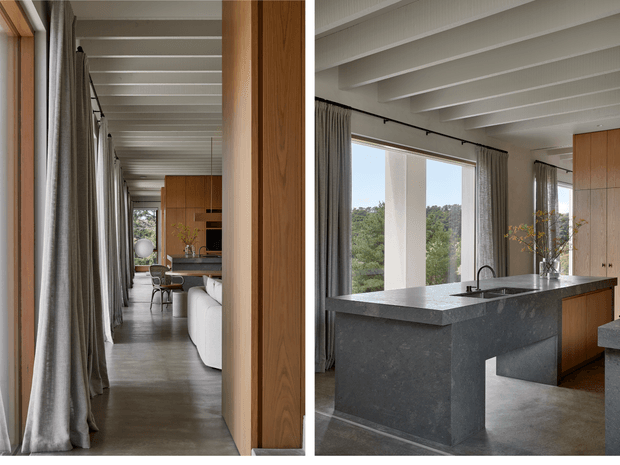 A refined open-plan interior showing a contemporary kitchen opening toward a long living and dining zone. The kitchen features a substantial dark stone island with integrated sink and tap, warm timber cabinetry, pale exposed ceiling beams, full-height windows with grey curtains, and a polished concrete floor. A corridor-like view extends toward dining furniture and a partially visible sofa, with greenery visible outside.