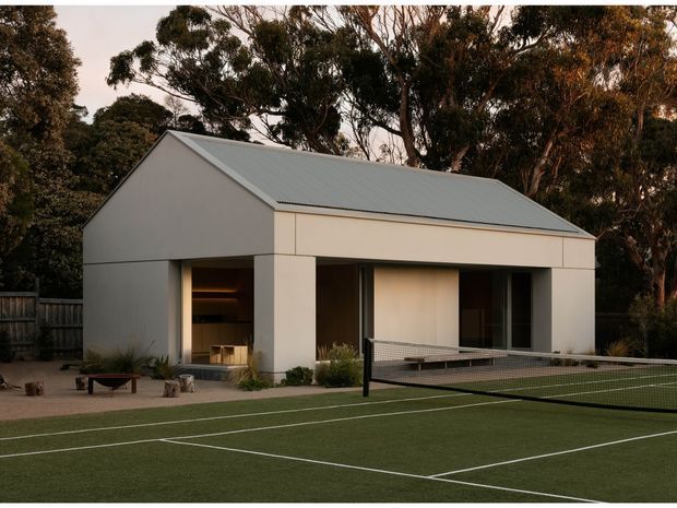 A minimalist single-storey pavilion-like building beside a private tennis court. The structure has a simple gabled form, pale rendered walls, a light grey metal roof, broad recessed openings, and warm interior lighting, set among mature trees and outdoor landscaping.
