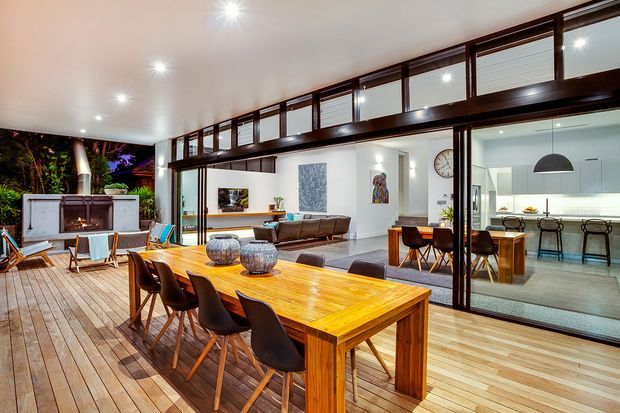 A spacious open-plan indoor-outdoor entertaining area with a large timber dining table and chairs on a timber deck, opening through extensive dark-framed glazing to a contemporary living room and white kitchen. The space includes an outdoor fireplace, lounge seating, pendant lighting, and high clerestory-style windows beneath a broad ceiling.
