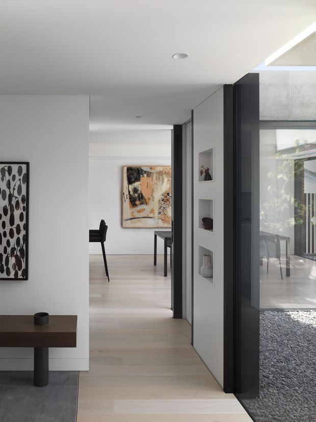 A minimalist contemporary residential interior showing a bright hallway or circulation space opening toward a dining area. Pale timber flooring, white walls and ceiling, dark charcoal door and window frames, recessed wall niches, and large glazed openings create a clean, gallery-like composition. Abstract artwork, a slim dining table and chairs, and a dark timber console are visible. A gravel-covered exterior courtyard is seen through the glazing on the right.