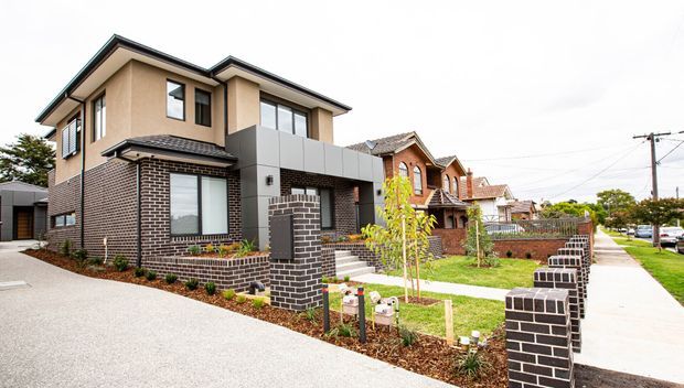 Street-facing view of a newly built multi-storey contemporary residence with a dark brick lower level, rendered upper walls, charcoal roof edges, and grey metal-clad upper facade elements. The property includes a paved driveway, front garden, pedestrian path, and low brick boundary fencing, with neighbouring suburban homes visible beyond.