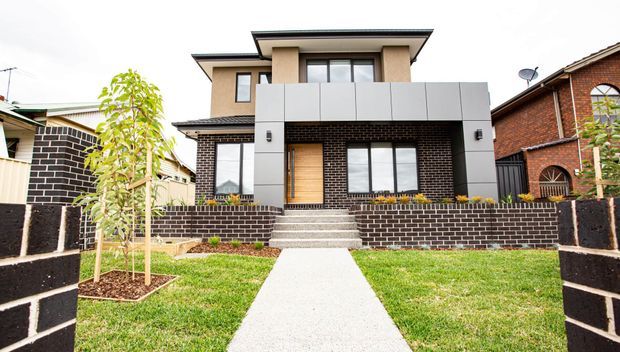Front exterior view of a contemporary multi-storey suburban house with dark brickwork, light rendered upper walls, a projecting box-like upper facade, timber front door, broad concrete steps, and a straight concrete path through a landscaped lawn. Adjacent brick homes are visible on either side.