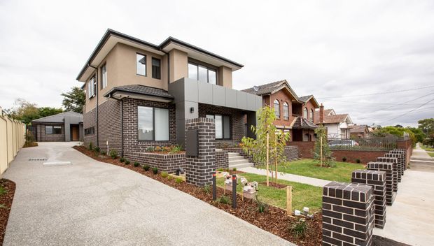 Exterior view of a contemporary multi-storey brick residence in a suburban streetscape. The home combines dark brickwork, rendered upper walls, a projecting light-grey cladding element, dark-framed windows, a driveway, landscaped front garden, and low brick boundary fencing. Adjacent traditional brick homes are visible along the street.