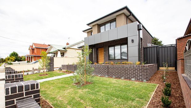 Front exterior view of a contemporary two-storey suburban home with dark brickwork, light grey upper cladding, black-framed glazing, a shallow pitched roof, and a landscaped front yard. A low brick retaining wall, paved entry path, fencing, and neighbouring homes are visible.