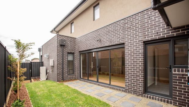Exterior view of a contemporary multi-level residential building with dark brick lower walls, beige rendered upper walls, dark-framed glazing, and a small landscaped courtyard. A paved path of square stepping stones leads to large sliding glass doors and a secondary entry, with fencing and outdoor HVAC equipment visible.