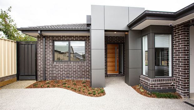 Front exterior view of a contemporary single-storey brick home with a low-pitched roof, charcoal metal cladding accents, timber entry door, projecting bay window, concrete paths, and a timber privacy fence.