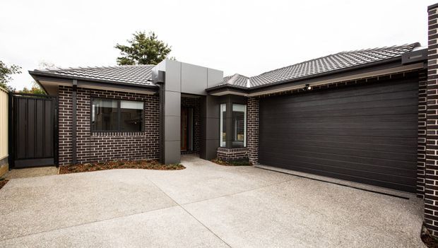 Single-storey residential facade viewed from the driveway, featuring dark brown brick walls with light mortar, grey tiled hipped roof sections, a charcoal double garage door, dark metal entry and side gates, and a pale textured concrete driveway. The design is compact and contemporary with traditional pitched roof forms and strong horizontal brickwork.