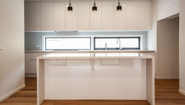 Bright contemporary kitchen with a large white island, glossy white full-height cabinetry, a grey splashback, timber flooring, horizontal windows above the sink, and three black pendant lights.