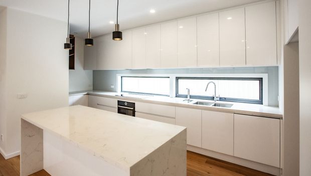 Bright contemporary kitchen with a large white stone island, extensive glossy white overhead cabinetry, pale lower cabinets, a long horizontal window above the sink, dark pendant lights, and warm timber flooring.