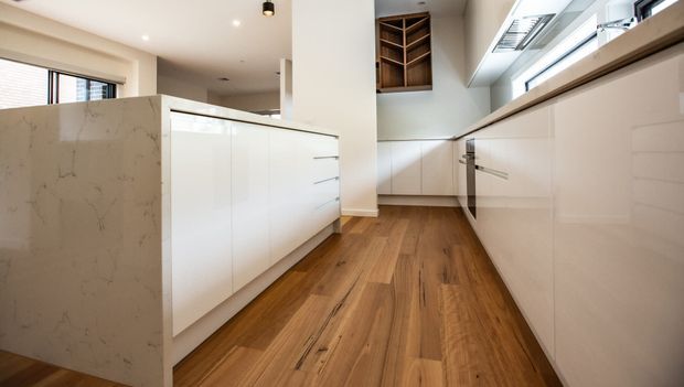 Bright contemporary kitchen featuring white flat-panel cabinetry, a pale veined stone island, warm timber flooring, integrated appliances, and a high horizontal window providing abundant natural light. An open timber display cabinet adds contrast above the rear cabinetry.