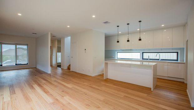 Bright open-plan interior showing a contemporary kitchen and adjoining living area. The kitchen has white flat-panel cabinetry, a pale stone island benchtop, a long horizontal window above the benchtop, and three black pendant lights. The adjoining room has warm timber flooring, white walls, recessed ceiling lights, and large windows.