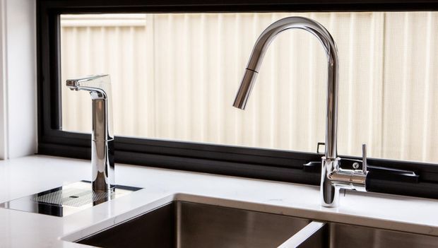 Close-up view of a contemporary kitchen sink area with two polished chrome faucets, a deep stainless-steel double-bowl sink, white benchtops, and a large black-framed window with sheer blinds.