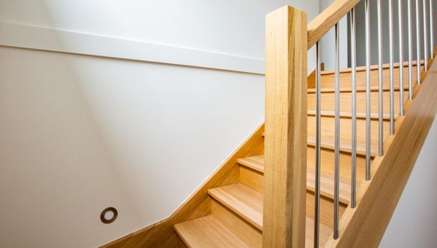 Close-up interior view of a modern timber staircase with light wood treads, stringers, posts and handrails, paired with slender dark metal balusters. The stairwell has smooth off-white walls, a simple white ceiling trim, and a small recessed wall light.