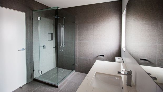 Contemporary bathroom featuring a frameless glass walk-in shower, large dark grey wall and floor tiles, a white floating vanity with an integrated basin, chrome tapware, and a large wall mirror. The space has a clean, refined, minimalist appearance.